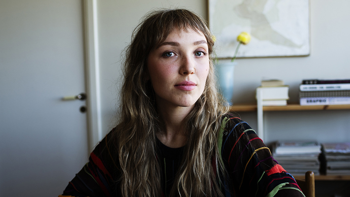 Ung kvinna som sitter i hemmiljö och tittar in i kameran.